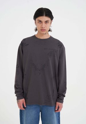 Junge Person mit mittellangen dunklen Haaren, die ein dunkelgraues Langarmshirt mit Totenkopf- und Flügeldesigns und blaue Jeans trägt.