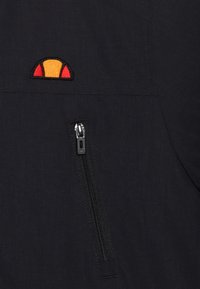 Veste en tissu noir avec une poche zippée. Logo brodé orange et jaune remarquable sur la poitrine, avec une finition texturée.