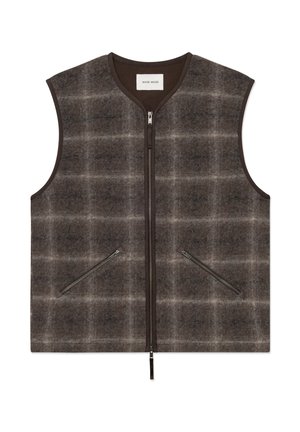 Jeune homme portant une casquette noire, un gilet à carreaux marron, une chemise rayée foncée, un pantalon noir et des chaussures noires, les mains dans les poches du gilet.