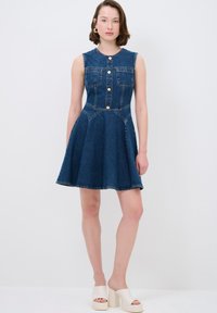 Robe en denim avec un corsage ajusté, fermeture à boutons et deux poches sur la poitrine. Jupe évasée, couleur bleue et coutures contrastées. Sandales à talons blocs blanches.