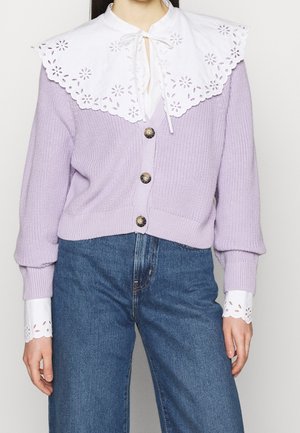 Femme portant un cardigan en tricot lavande avec un grand col en broderie anglaise blanche et des poignets assortis, associé à un jean bleu taille haute.