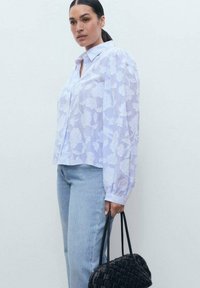 Blouse lilas clair avec un motif floral blanc, coupe décontractée, col boutonné et manches longues. Associée à un jean bleu et un sac tissé noir.