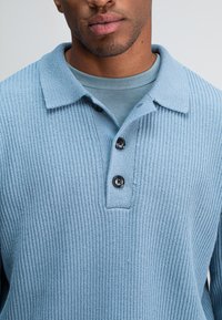 Hellblaues, geripptes Strickhemd mit Kragen, das mit drei schwarzen Knöpfen versehen ist und darunter ein geschichtetes hellblaues Hemd hat.