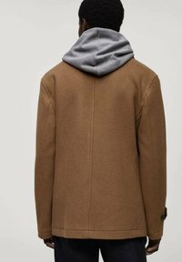 Bruine wolmix jas met een grijze hoodie eronder. Bijzondere details zijn een knoop op de mouw en een schone, gestructureerde vormgeving.