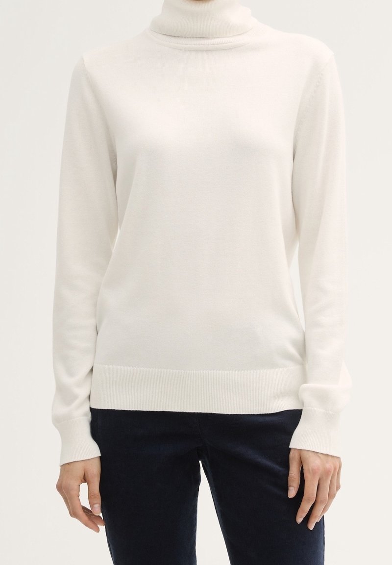 Bílý rolákový svetr s dlouhým rukávem nošený přes tmavé kalhoty, ruce volně podél těla na jednoduchém světlejší pozadí.