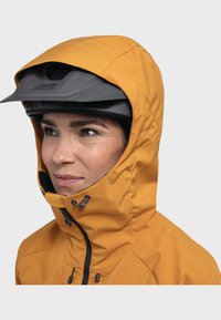 Gelbe wasserdichte Jacke mit hohem Kragen, verstellbarem Kapuze und schwarzem Reißverschluss. Unter der Kapuze wird ein schwarer Helm getragen.