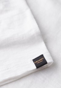 Dichtbij shot van witte stof met een textuurafwerking, voorzien van een zwarte label met gouden tekst die "SUPERDRY PREMIUM" zegt.