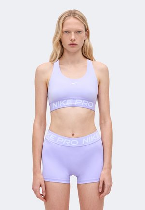Un mannequin se tient face à l’objectif, portant une brassière de sport Nike couleur lavande et un short de cyclisme assorti, devant un fond clair uni.