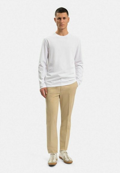 Hvid langærmet skjorte, beige bukser og hvide sneakers med beige accenter. Afslappet pasform, glat stof og minimale designdetaljer.