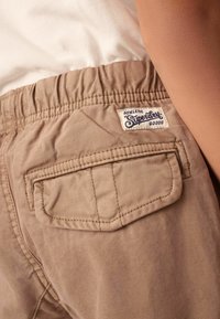 Beige byxors med elastisk midja som har en bakficka med en sömmad kant och en vävd etikett som säger "Superdry Athletic Goods."
