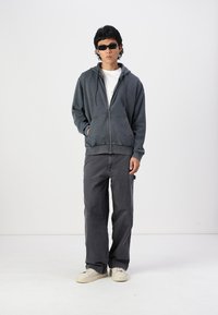 Gray zip-up hoodie with a front pocket, paired with loose gray pants and white sneakers. Model wears dark sunglasses and a white t-shirt.