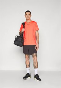 Bright orange short-sleeve shirt with logo, black shorts, and black sneakers with green accents. Model holding a black bag, showing tattoos.