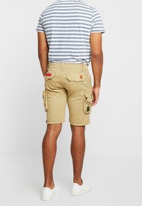 Mężczyzna w beżowych szortach cargo i szaro-białej w paski koszulce, stoi plecami do kamery na białym tle.