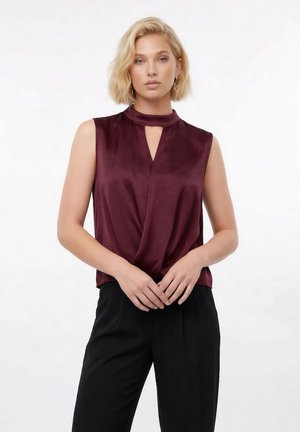 Blond kvinne har på seg ermeløs, mørk burgunder satengbluse med nøkkelhullutringning og vide svarte bukser, stående mot hvit bakgrunn.