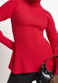 Červený žebrovaný rolák s dlouhými rukávy a rozšířenou siluetou; vyroben z texturované pleteniny. Obsahuje boční rozparky pro větší detail.