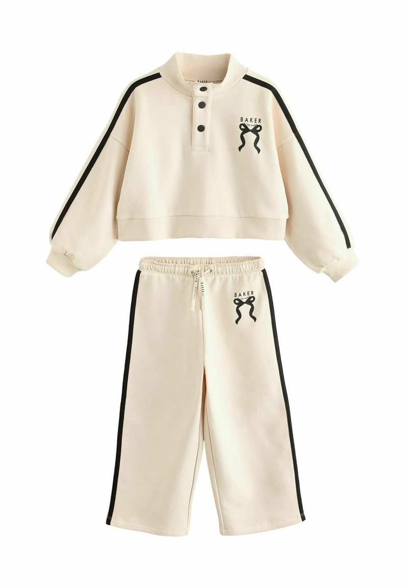 Survêtement couleur crème comprenant un pull court avec des boutons noirs et un logo, associé à un pantalon ample avec des bandes latérales.