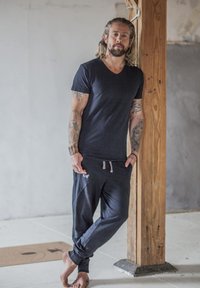 T-paita merivärissä, V-kaula-aukolla, ja siihen sopivat jogger-housut, joissa on vaalea nauha. Materiaalin näyttää olevan pehmeä; kuvassa on tatuointeja ja paljaat jalat. Taustalla puupylväs.