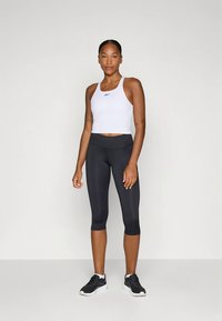 Witte cropped top met logo, zwarte capri-legging en zwarte sportschoenen. De outfit is nauwsluitend en gemaakt van gladde materialen.