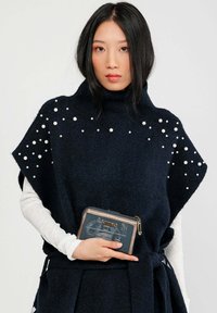 Poncho knit blu navy con colletto alto, impreziosito da perle bianche. Abbinato a un portafoglio blu scuro con chiusura a zip e design grafico.