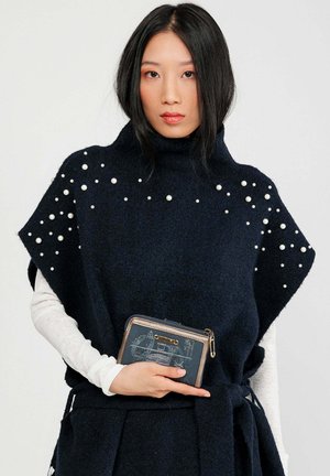 Marinefarbener Strickponcho mit hohem Kragen, verziert mit weißen Perlen. Kombiniert mit einer dunkelblauen Geldbörse mit Reißverschluss und grafischem Design.