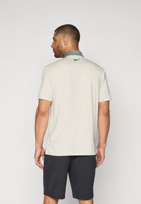 Vīriešu īsām piedurknēm polo krekls gaiši bēšā krāsā ar tumši zaļu apkakli un mazu melnu Nike logotipu aizmugurē; valkāts ar tumšām šortiem.