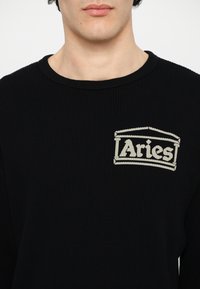 Sorte waffle-strikkede sweater med et kremfarvet broderet "Aries" logo på venstre side af brystet. Rund halsudskæring og struktureret stof.