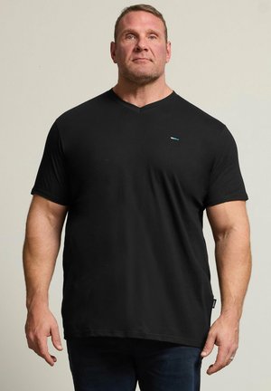 Muskeløs mand står mod en ensfarvet baggrund iført en ensfarvet sort kortærmet V-hals T-shirt og mørke bukser.