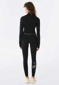 Černý sportovní top s dlouhým rukávem a legíny. Top má vysoký límec a bílý text na lemu. Legíny mají bílý grafický detail.
