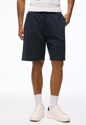 Man draagt marineblauwe short met trekkoord, witte sokken, witte sneakers en een wit mouwloos shirt met handen in de zakken.