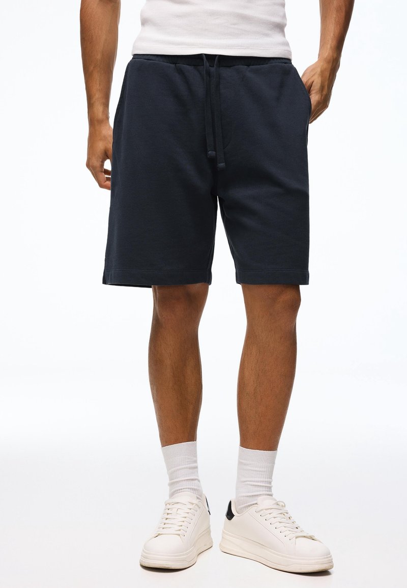 Homme portant un short bleu marine à cordon, des chaussettes blanches, des baskets blanches et un débardeur blanc, les mains dans les poches.