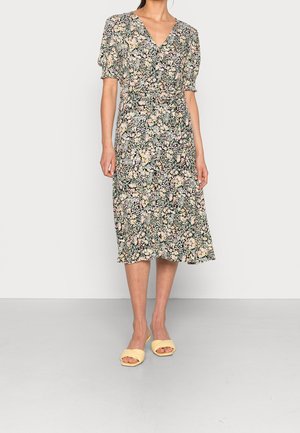 Femme portant une robe fleurie mi-longue avec des manches bouffantes et un décolleté en V, associée à des talons ouverts jaunes clairs, debout sur un sol uni.