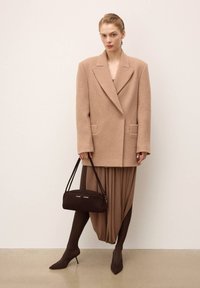 Beige wool blend oversized blazer with flap pockets, paired with a brown asymmetric pleated dress. Dark brown suede handbag and pointed heels.