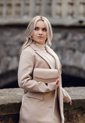 Beige leather crossbody bag with a zip closure, smooth texture, subtle logo detail, and a detachable strap, held by a person in a coat.