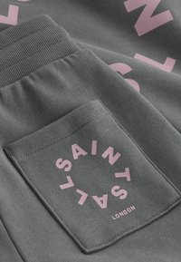 Sivé tkané s vreckom so stehovaním, na ktorom je ružový kruhový nápis "Saint Saults London" a časť veľkého ružového textu na odeve.