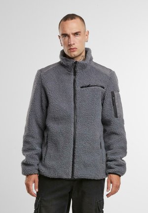 Veste en polaire grise avec un col montant, une fermeture éclair sur le devant et plusieurs poches, avec des accents gris sur les épaules. Surface texturée.