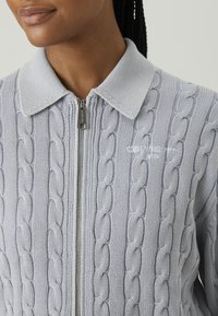 Light gray zip-up sweater featuring a cable-knit pattern, ribbed collar, and logo embroidery on the left side. Soft texture, relaxed fit.