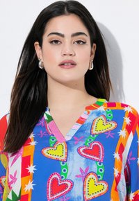 Blouse colorée et à motifs avec un col en V, des cœurs multicolores et un mélange de formes vives sur un fond bleu, fabriquée en tissu léger.