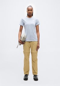 Γυναίκα που στέκεται μπροστά φοράει γκρι ανοιχτό t-shirt, μπεζ παντελόνι μετατρέψιμο, μαύρα παπούτσια πεζοπορίας και κρατά μια μπεζ τσάντα μέσης.