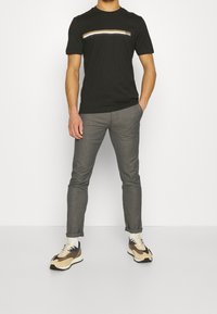 Černé triko s krátkým rukávem se pruhovaným vodorovným designem v hnědé, bílé a zlaté barv ě; šedé slim-fit kalhoty; béžové a hnědé tenisky.