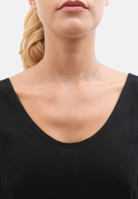 Collier choker en argent avec une chaîne délicate et un petit pendentif transparent, porté sur un cou nu, contre un haut noir en V.