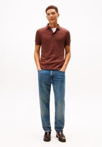 Joven de pie con las manos en los bolsillos, vistiendo una camiseta polo marrón, jeans azules y mocasines marrón oscuro, frente a un fondo blanco liso.