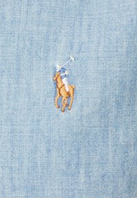 Ljust blått tyg med en liten broderad logotyp av en polo spelare på hästryggen i bruna och beige toner.