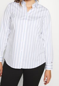 Lyseblå og hvide stribet skjorte med knapper, lange ærmer, buet kant og diskret logodetalje på brystet. Blødt bomuldsstof.