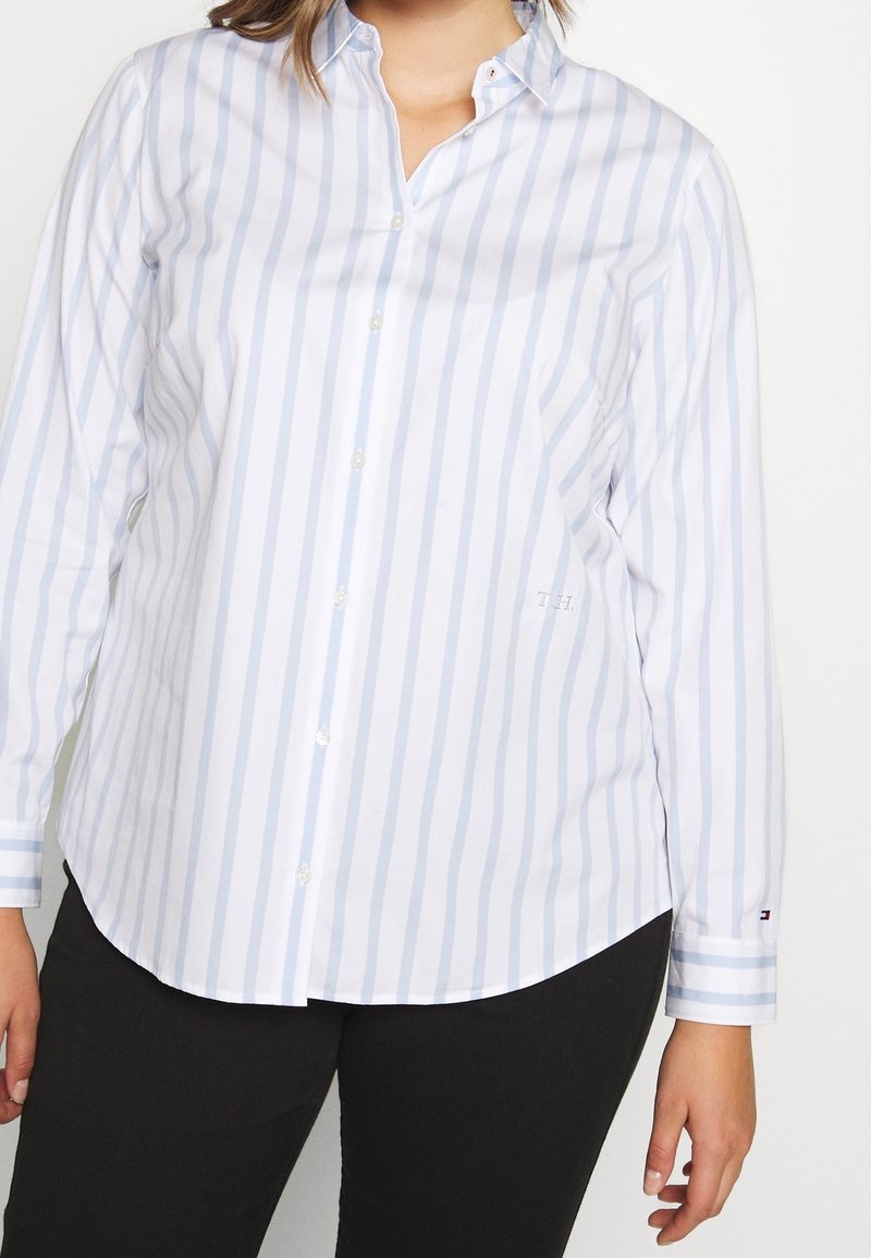 Lyseblå og hvide stribet skjorte med knapper, lange ærmer, buet kant og diskret logodetalje på brystet. Blødt bomuldsstof.