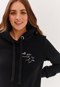 Černá mikina s kapucí a stahovací šňůrkou se stříbrnými detaily, na přední straně s bílým ručně psaným textovým designem. Hladká textura, volnější střih.
