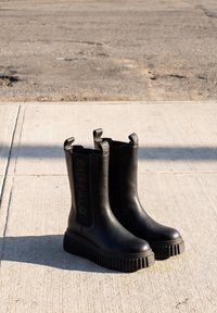 Paar schwarze Gummistiefel bis zur Mitte der Waden mit dicken, profilierten Sohlen, die auf einem sonnenbeschienenen Betonpflaster stehen.
