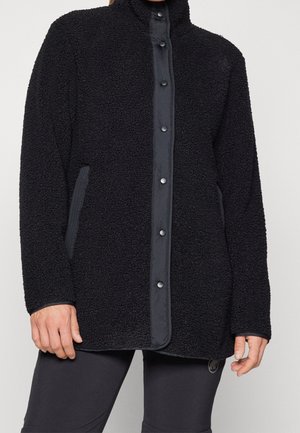 Veste en polaire noire texturée avec fermeture frontale à boutons-pression et poches latérales portée par une personne visible du cou jusqu'à la mi-cuisse.