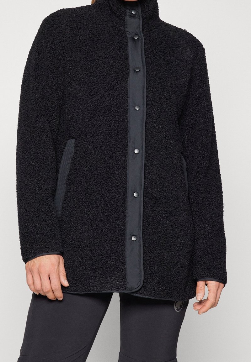 Veste en polaire noire texturée avec fermeture frontale à boutons-pression et poches latérales portée par une personne visible du cou jusqu'à la mi-cuisse.