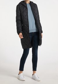 Zwarte gewatteerde lange jas met een capuchon, voorzien van een ritssluiting aan de voorkant, zijzakken en elastische boorden, gedragen over een lichtblauw sweatshirt en jeans.