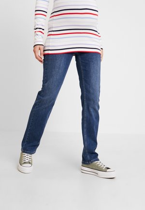 Osoba ubrana w niebieskie dżinsy, zielone tenisówki z materiału i koszulę z długim rękawem z białymi, czerwonymi, czarnymi i jasnoniebieskimi poziomymi paskami.